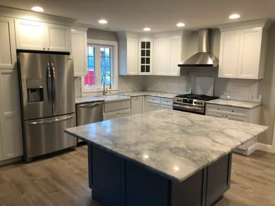 White shaker kitchen remodel with a marble-top island, stainless steel appliances, and a chimney range hood by KRidge Construction